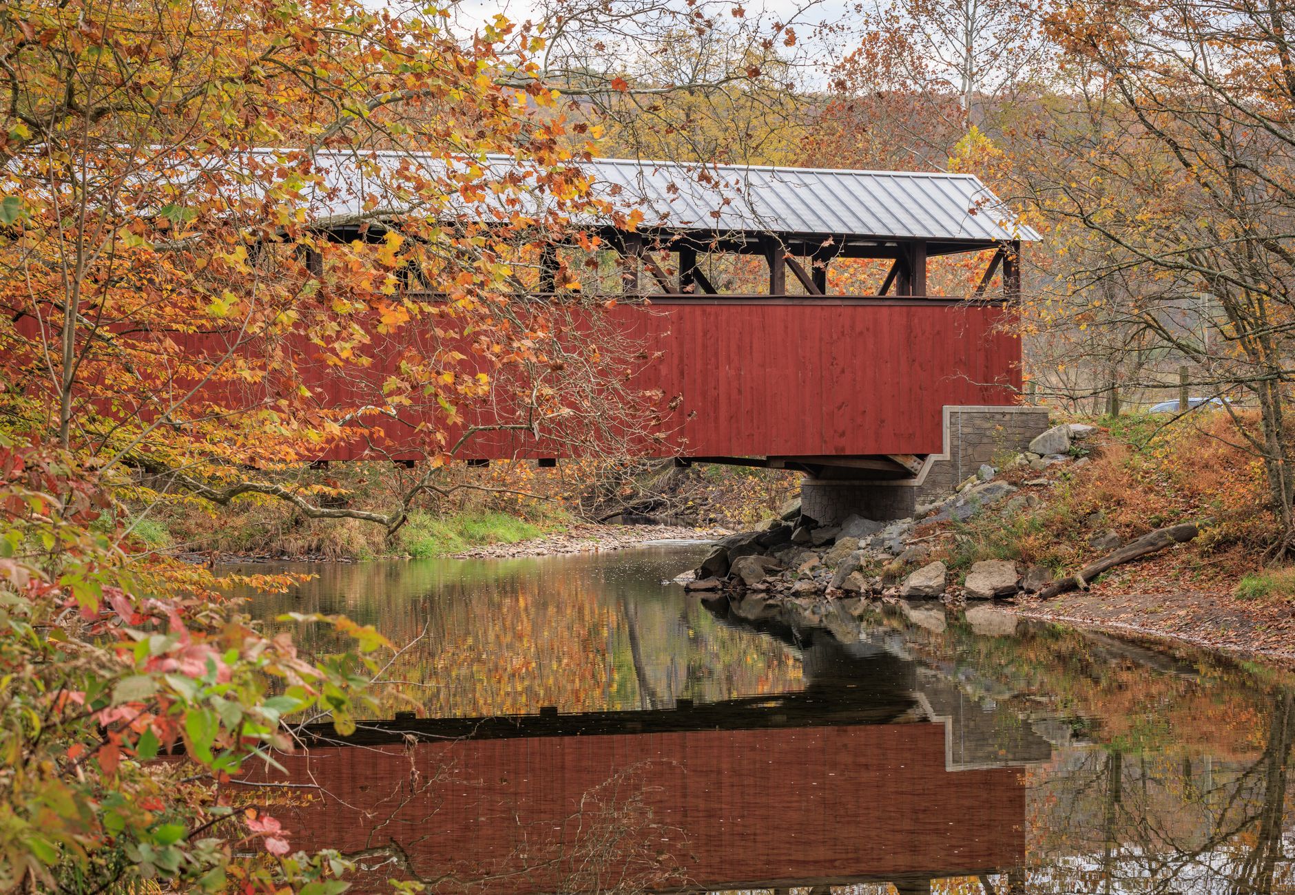 Beautiful autumn scene of a red covered bridge reflecting on a calm creek, surrounded by vibrant fall foliage in Pennsylvania.