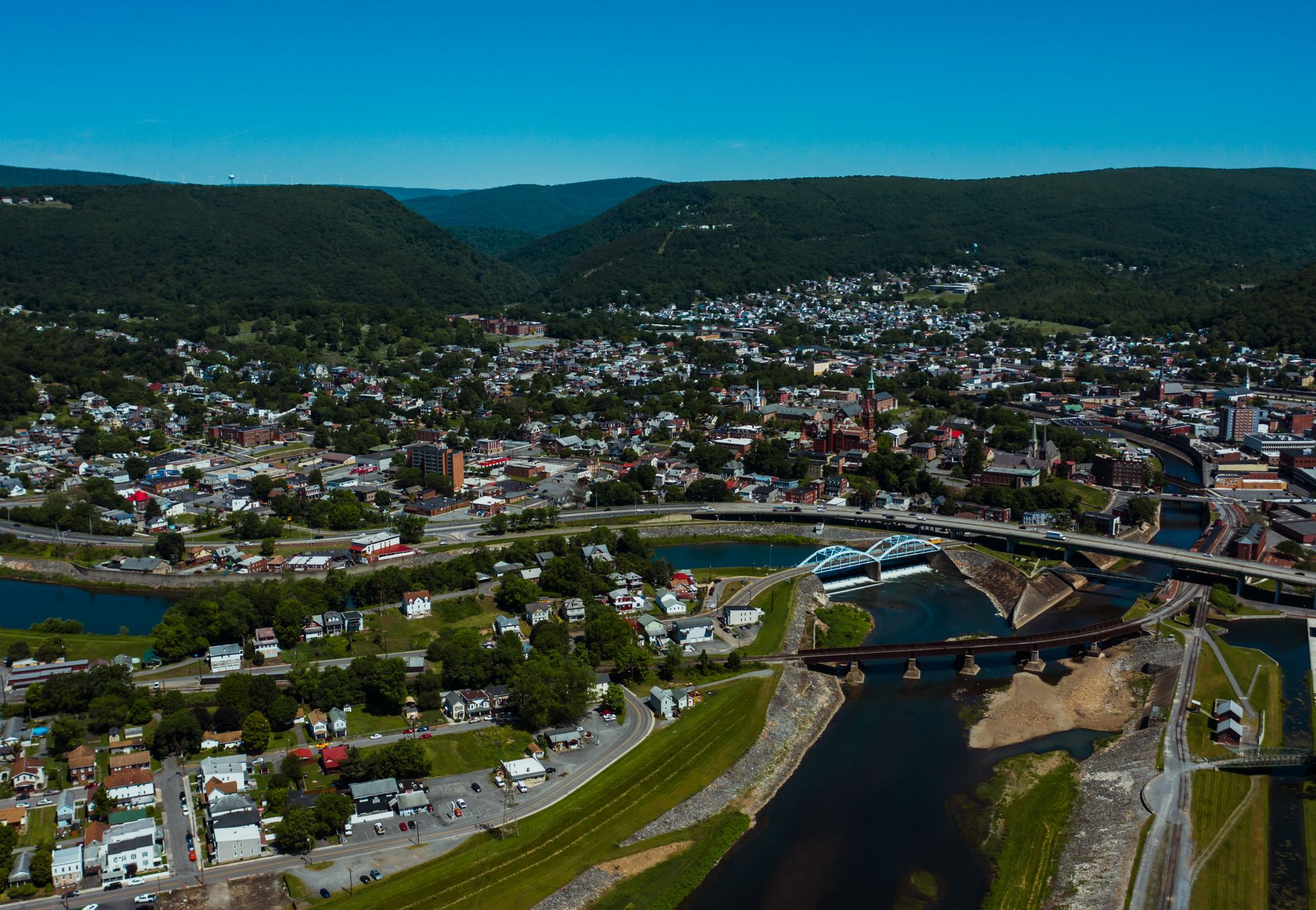 Aerial view of wavy roadway and bridges over canal between dwelling buildings near lawns with trees in mounts under blue sky